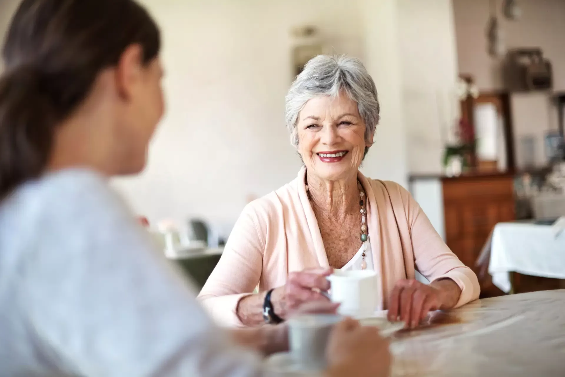 An elderly woman drinking tea and chatting with another woman.